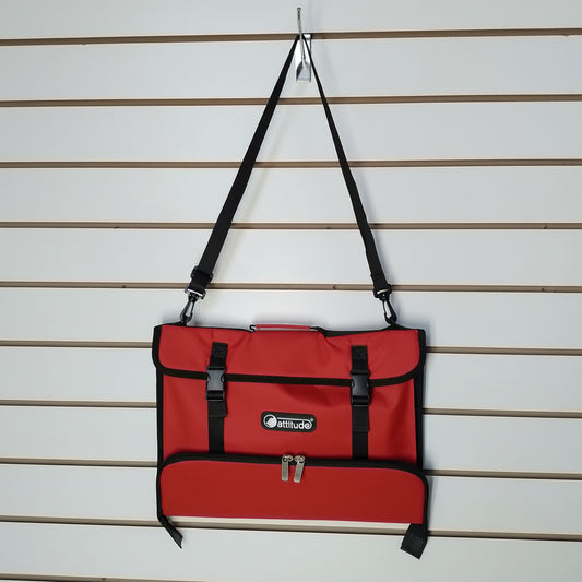 A red messenger bag with black straps and trim, hanging from a peg on a slatwall. The bag features a front pocket with buckle closures and a zippered lower pocket, with a visible 'Attitude' logo patch.