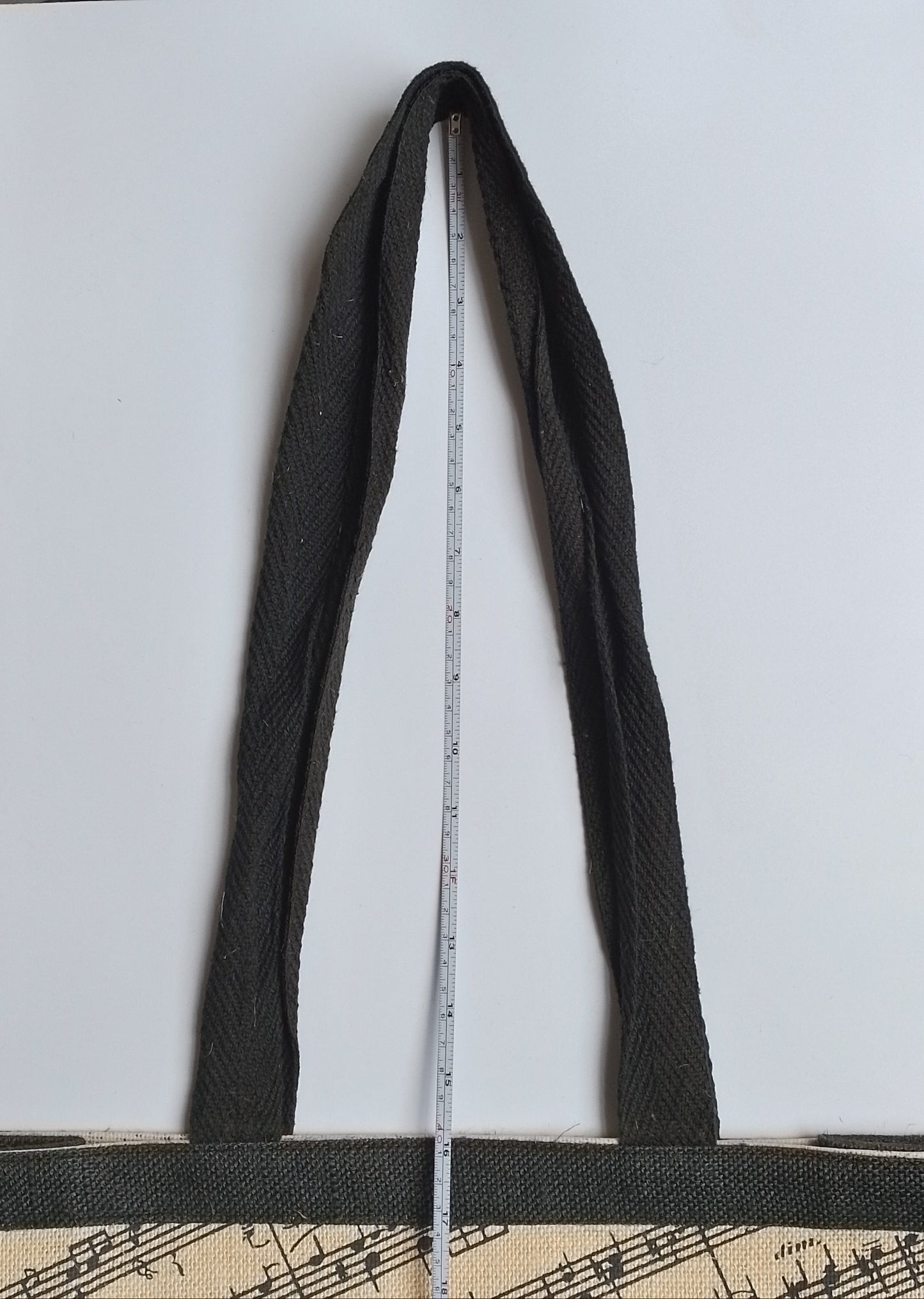 A black woven tote bag with a beige printed pattern at the bottom, shown with a metal tape measure hanging from its strap to indicate size, set against a plain white background.