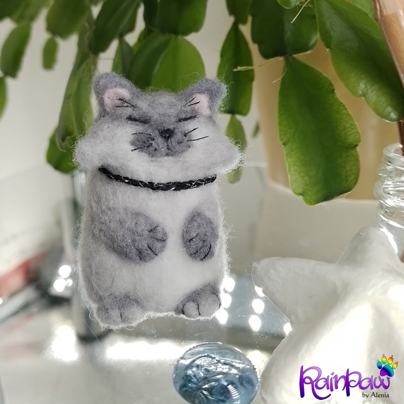 Felted gray cat figurine with a black collar on a reflective surface with green leaves in the background.