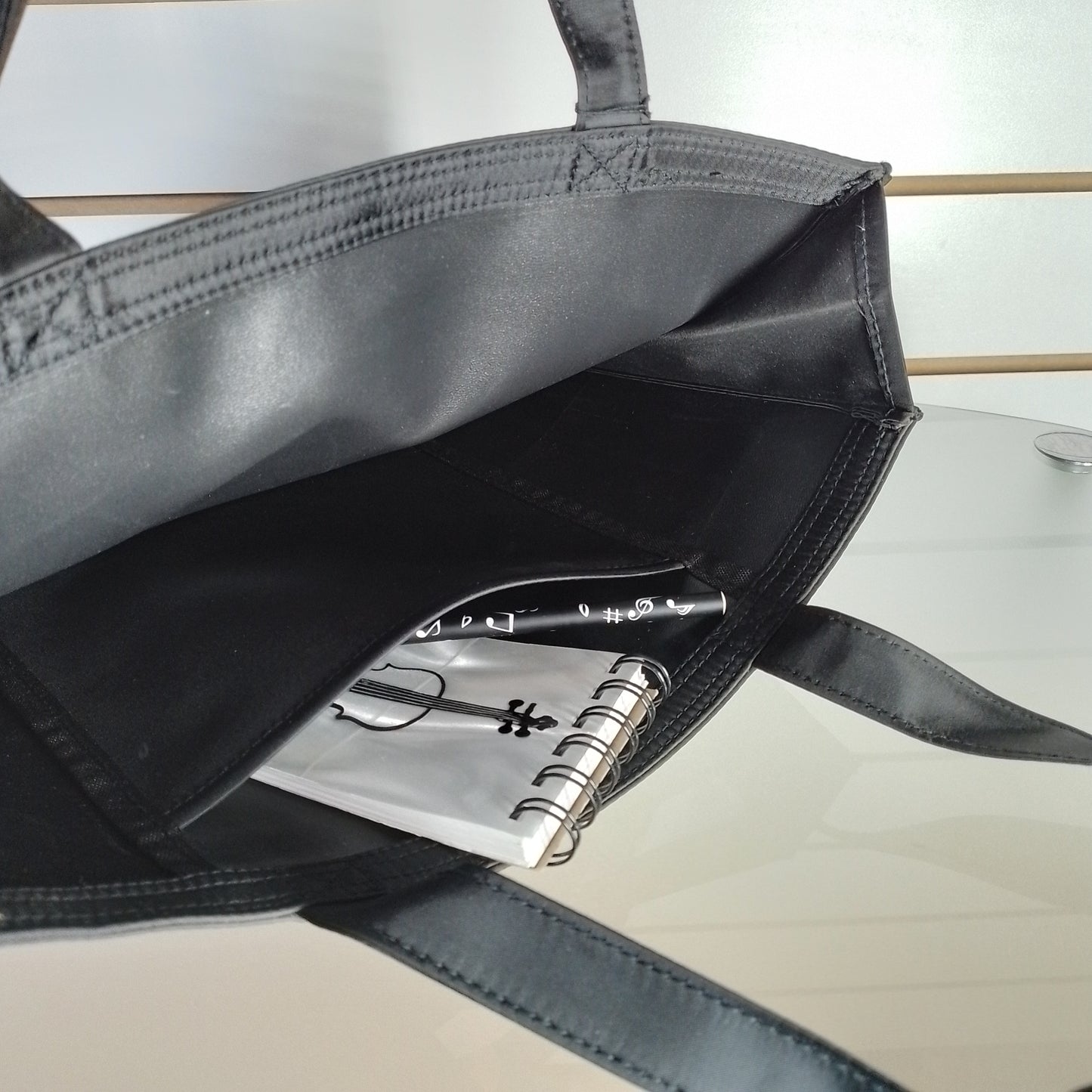 A black tote bag shown open, revealing a black interior pocket holding a small white spiral notebook and a black pen.