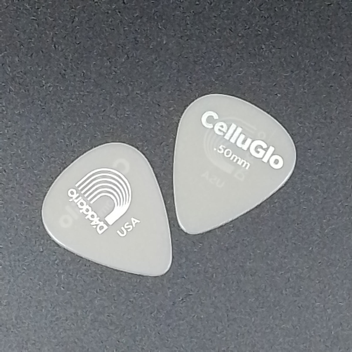 Two translucent CelluGlo 50mm guitar picks on a dark gray textured background, one showing the CelluGlo branding and the other showing the D'Addario USA logo.