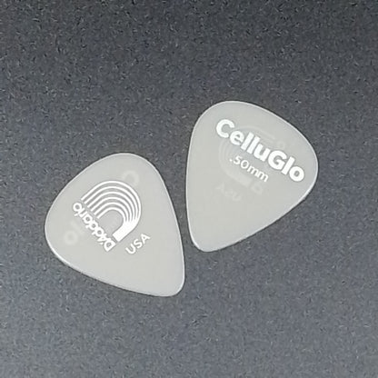 Two translucent CelluGlo 50mm guitar picks on a dark gray textured background, one showing the CelluGlo branding and the other showing the D'Addario USA logo.