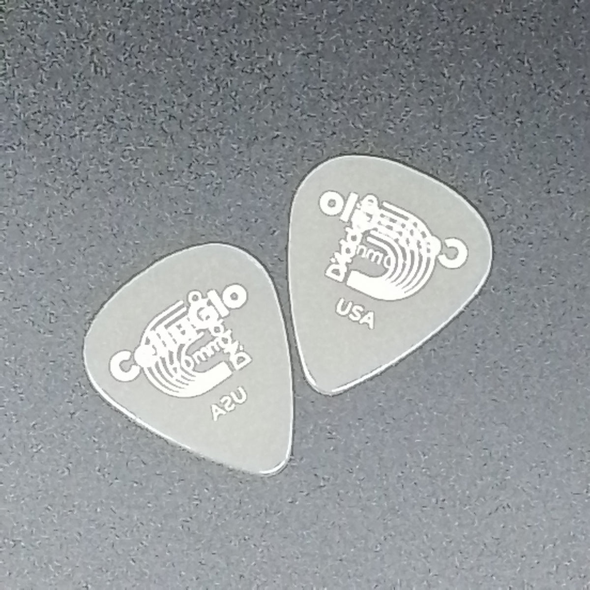 Two translucent gray guitar picks with white printed graphics and text, including 'C' and 'USA', placed on a dark gray textured surface.