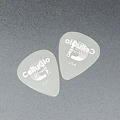 Two translucent gray guitar picks with white printed graphics and text, including 'C' and 'USA', placed on a dark gray textured surface.