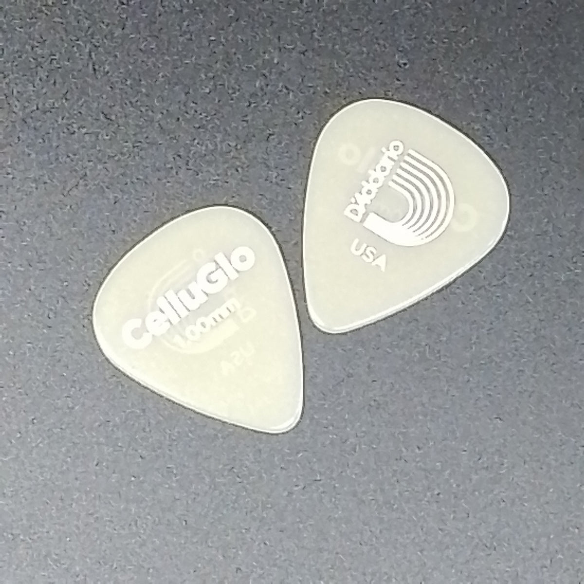 Two translucent plastic guitar picks on a dark gray textured background, one printed with the D'Addario logo and the other with the Calliope logo.