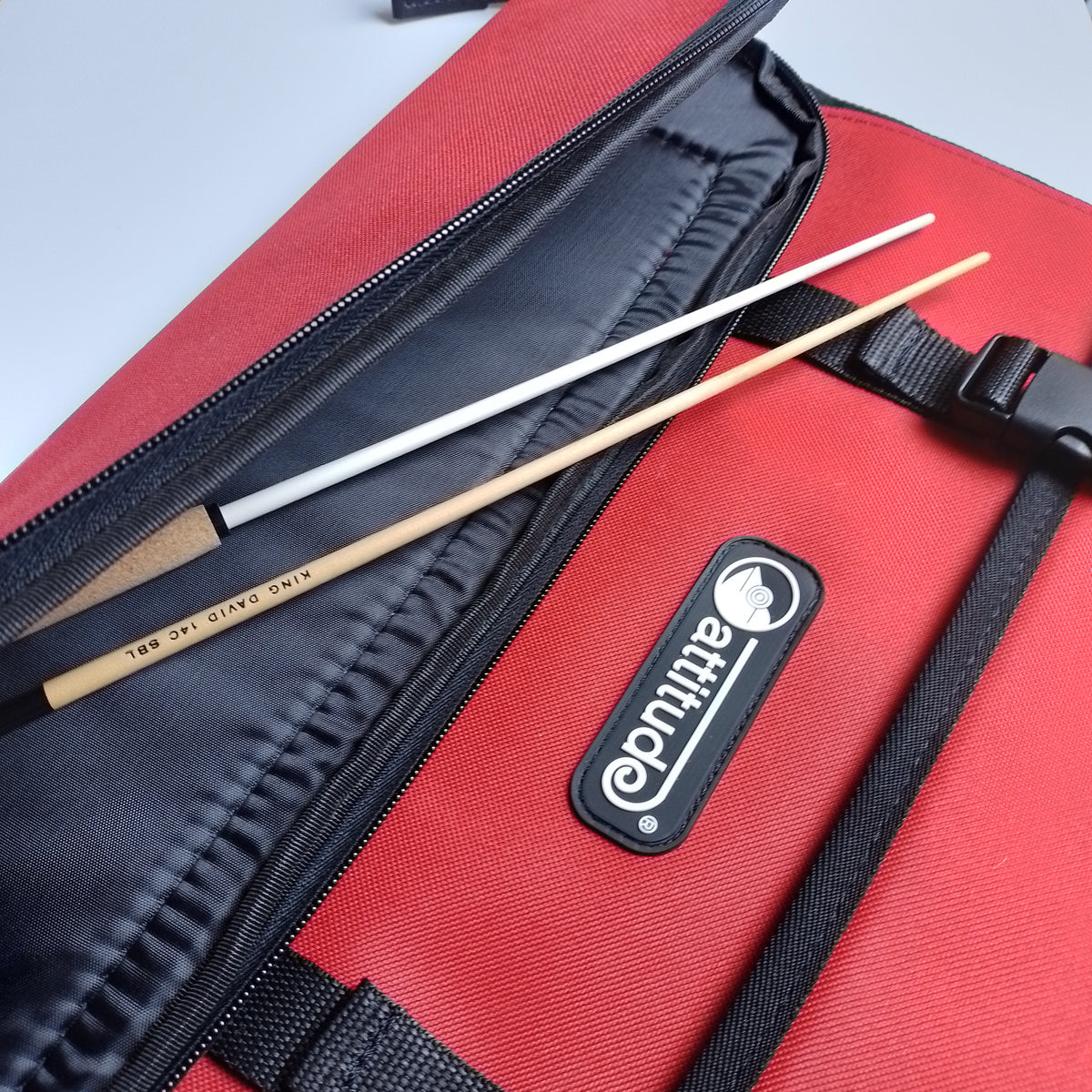A close-up of a red and black Attitude brand bag with a black zipper, showing two wooden batons resting inside.