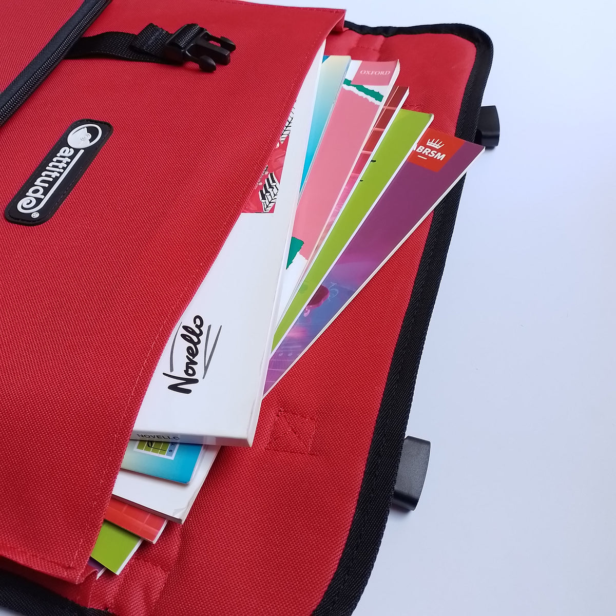A red fabric organizer pouch or document holder with black trim, shown open with colourful paper sheets and notebooks inside. A black oval brand patch on the front reads 'Attitude' and a notebook visible inside reads 'Norello'.
