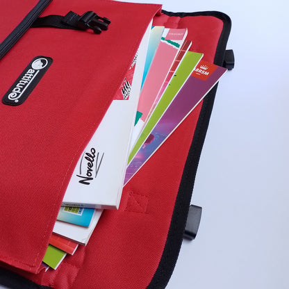 A red fabric organizer pouch or document holder with black trim, shown open with colourful paper sheets and notebooks inside. A black oval brand patch on the front reads 'Attitude' and a notebook visible inside reads 'Norello'.