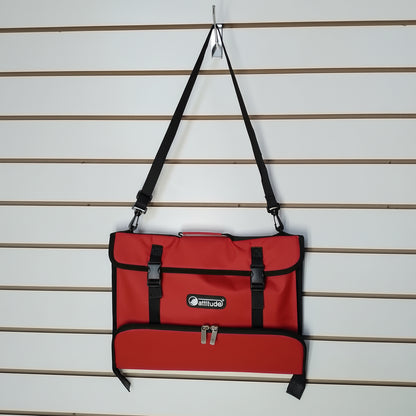 A red messenger bag with black straps and trim, hanging from a peg on a slatwall. The bag features a front pocket with buckle closures and a zippered lower pocket, with a visible 'Attitude' logo patch.