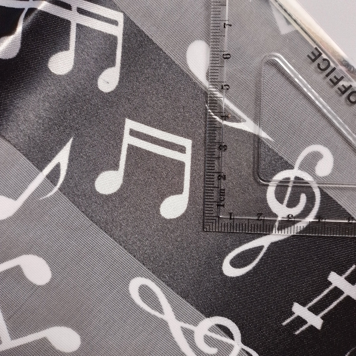 A close-up of a sheer black fabric with a white music note and clef pattern, laid over, a clear plastic ruler for proportion.