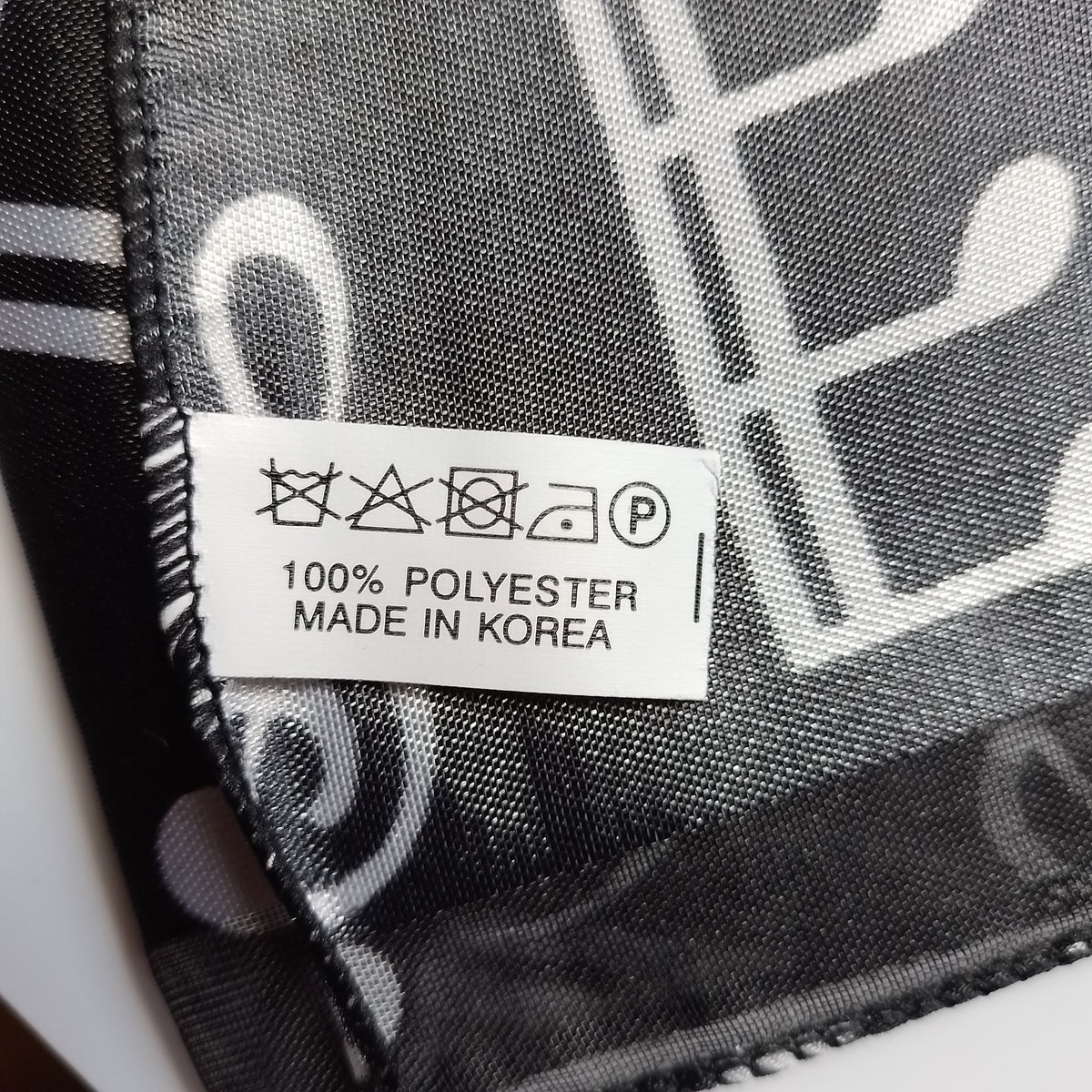 Close-up of a black and white patterned scarf with a white sewn-in care label reading '100% POLYESTER MADE IN KOREA'.