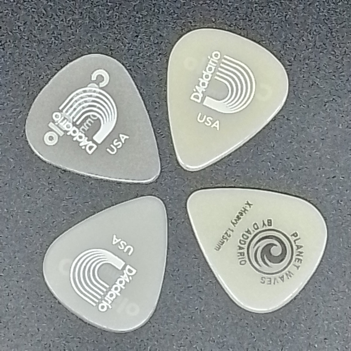 Four light lime green plastic guitar picks arranged in a square on a dark textured background. Each pick is printed with the D'Addario USA logo and one pick also shows Planet Waves by D'Addario text.