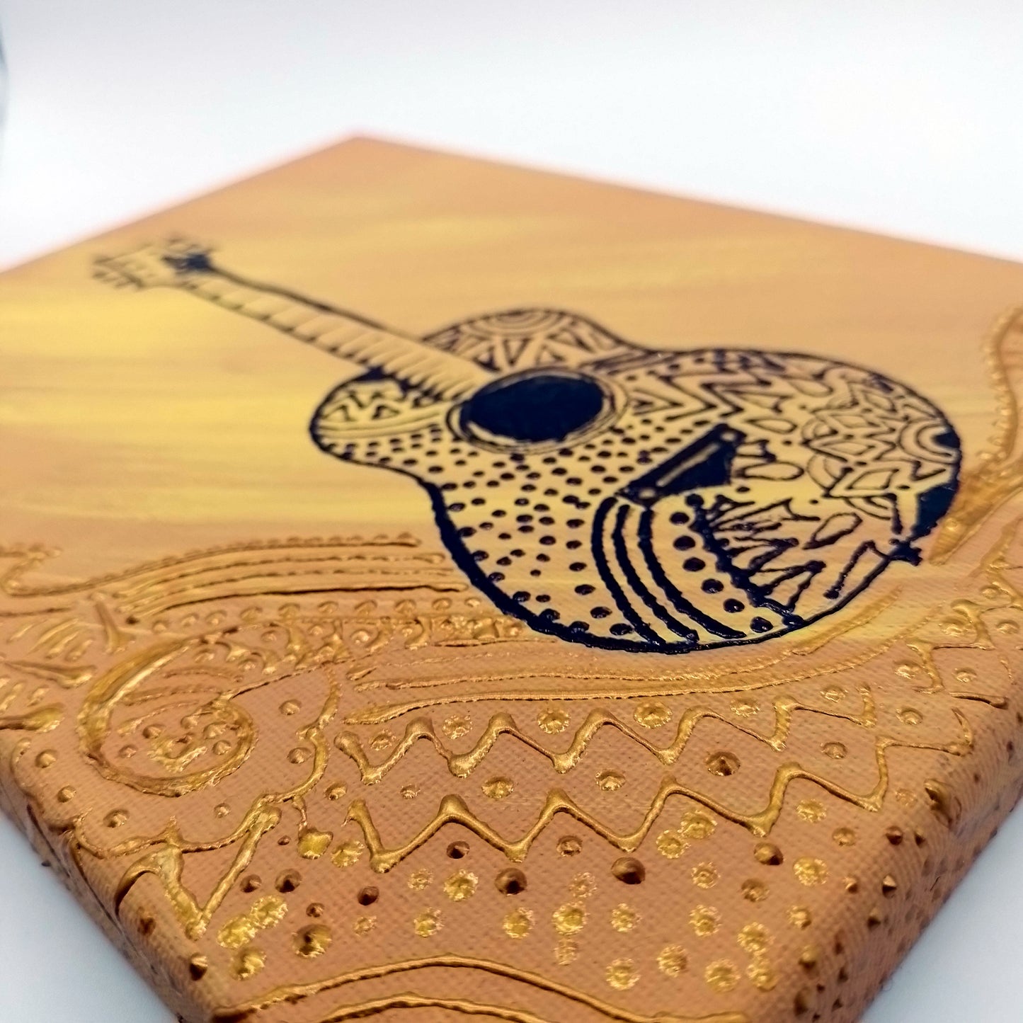 Detail  of the painting with black acoustic guitar on two tone orange and gold detail. The shape of the instrument is a light relief on a flat surface. 