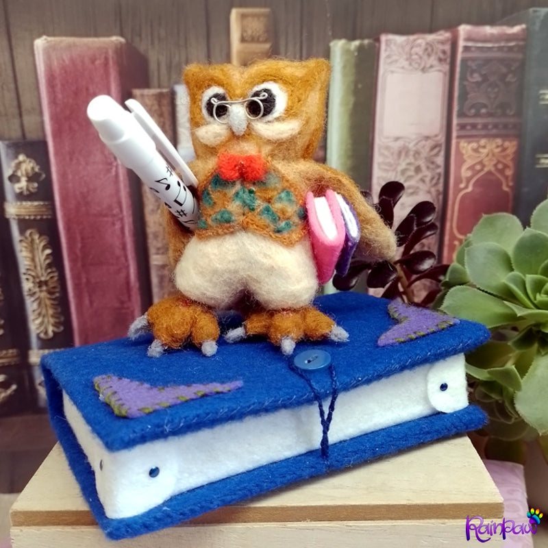A felted wool owl with a felt book shaped box and stationery items on a wooden surface with books in the background.