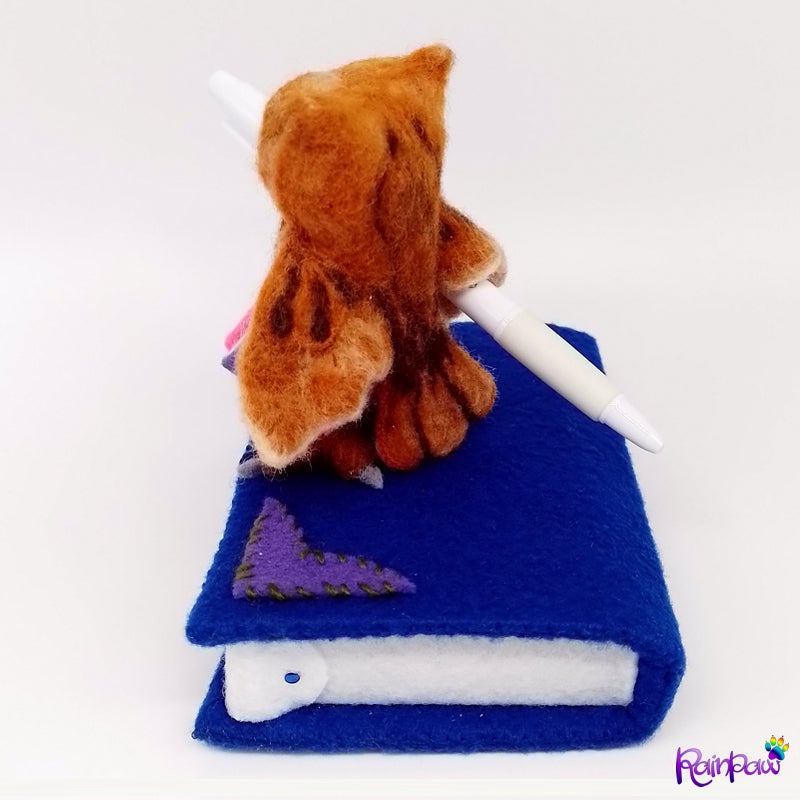 Back view of an owl on top of a blue book with purple decorative details. The owl serves as a pen holder and has a functioning pen under its wing