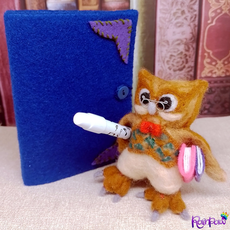 A felted wool owl with a felt book shaped box and stationery items on a wooden surface with books in the background.