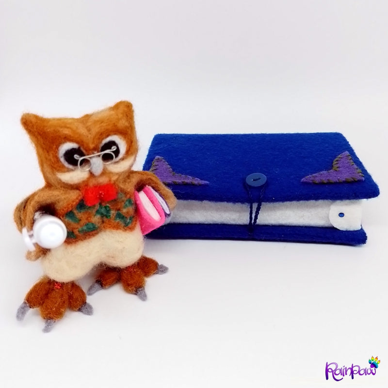 A felted wool owl with a felt book shaped box and stationery items on a wooden surface with books in the background.