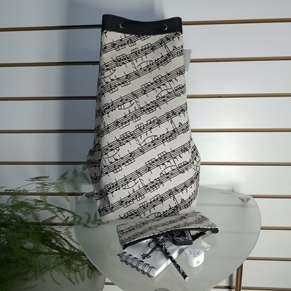 A drawstring bag with a black and white musical staff pattern, displayed on a glass table with a matching pouch, notebook, and pen.