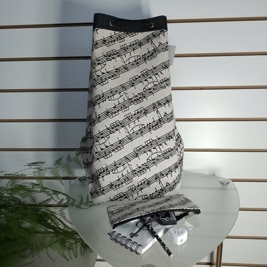 A drawstring bag with a black and white musical staff pattern, displayed on a glass table with a matching pouch, notebook, and pen.