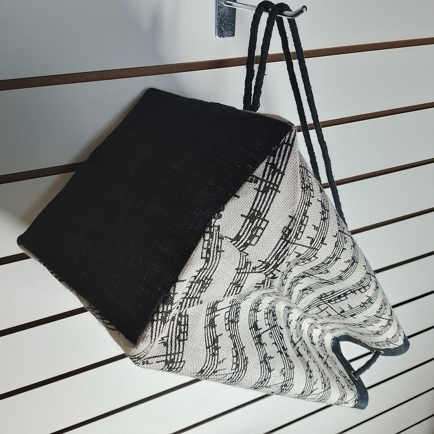 A fabric drawstring bag with a black and white musical note pattern on the upper half and a solid black lower half, hanging upside-down against a white slatwall.