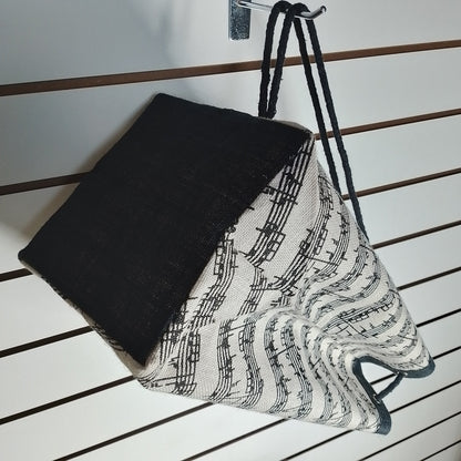 A fabric drawstring bag with a black and white musical note pattern on the upper half and a solid black lower half, hanging upside-down against a white slatwall.