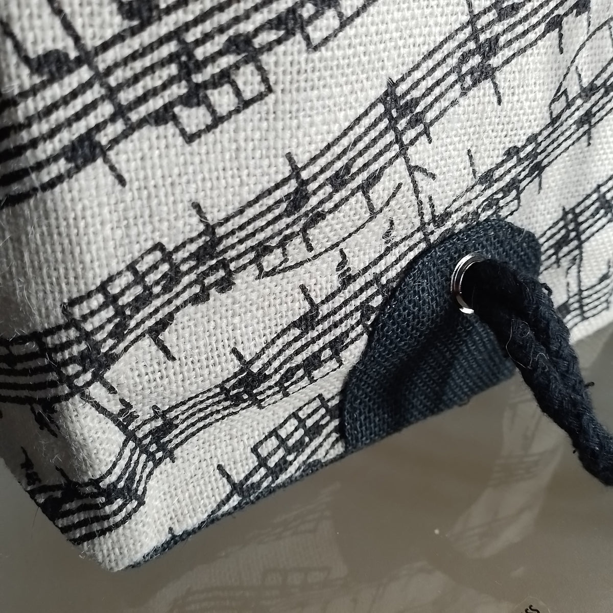 Close-up of a canvas bag featuring a black music staff and notes print on a white background, with a dark blue fabric accent and a braided black strap threaded through a metal grommet.
