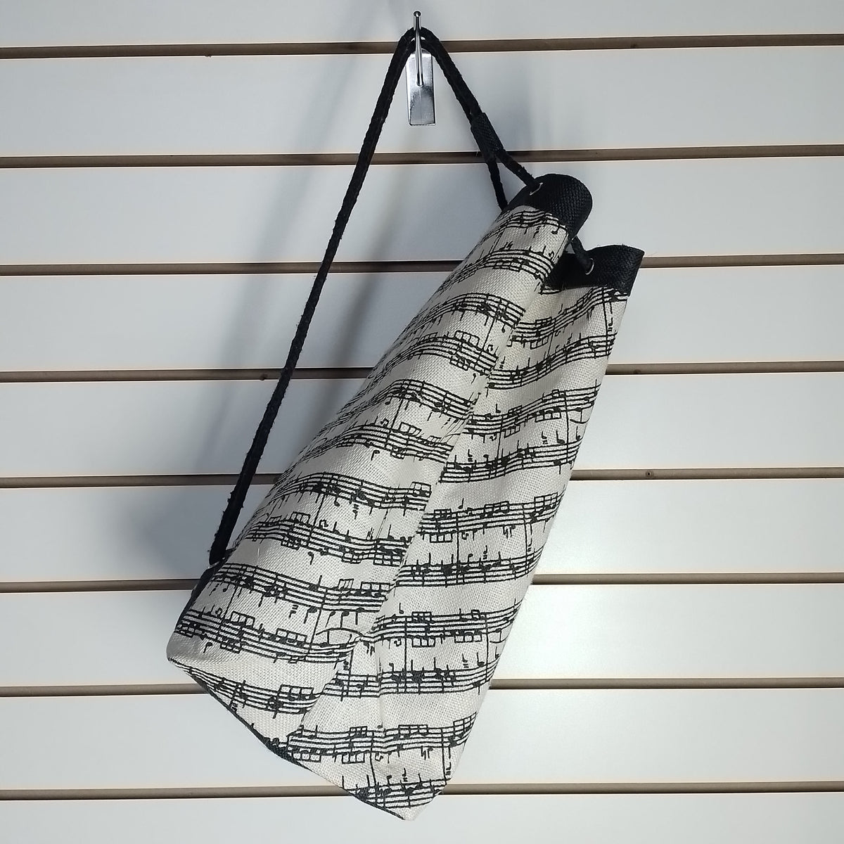 A fabric tote bag with a black and white musical sheet music pattern, hanging from a black strap on a white slatwall.