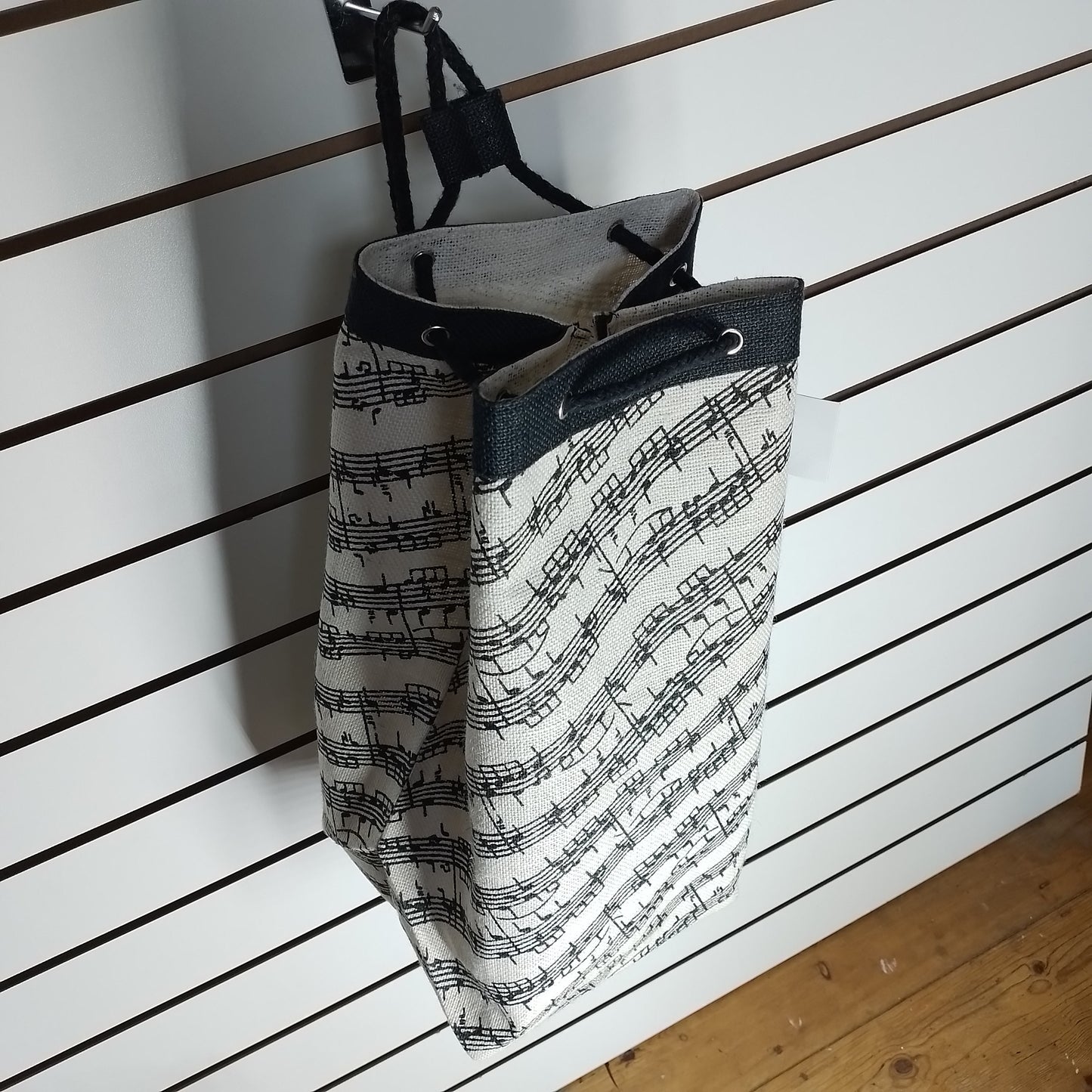 A fabric wall-hanging tote bag with a black and white musical sheet music pattern, hanging from a black hook on a white slatwall.