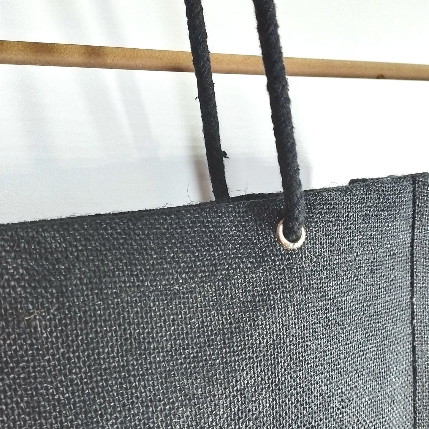 Close-up of a black canvas or heavy fabric panel with a metal grommet and black cord, shown against a white background with part of a wooden pole visible.