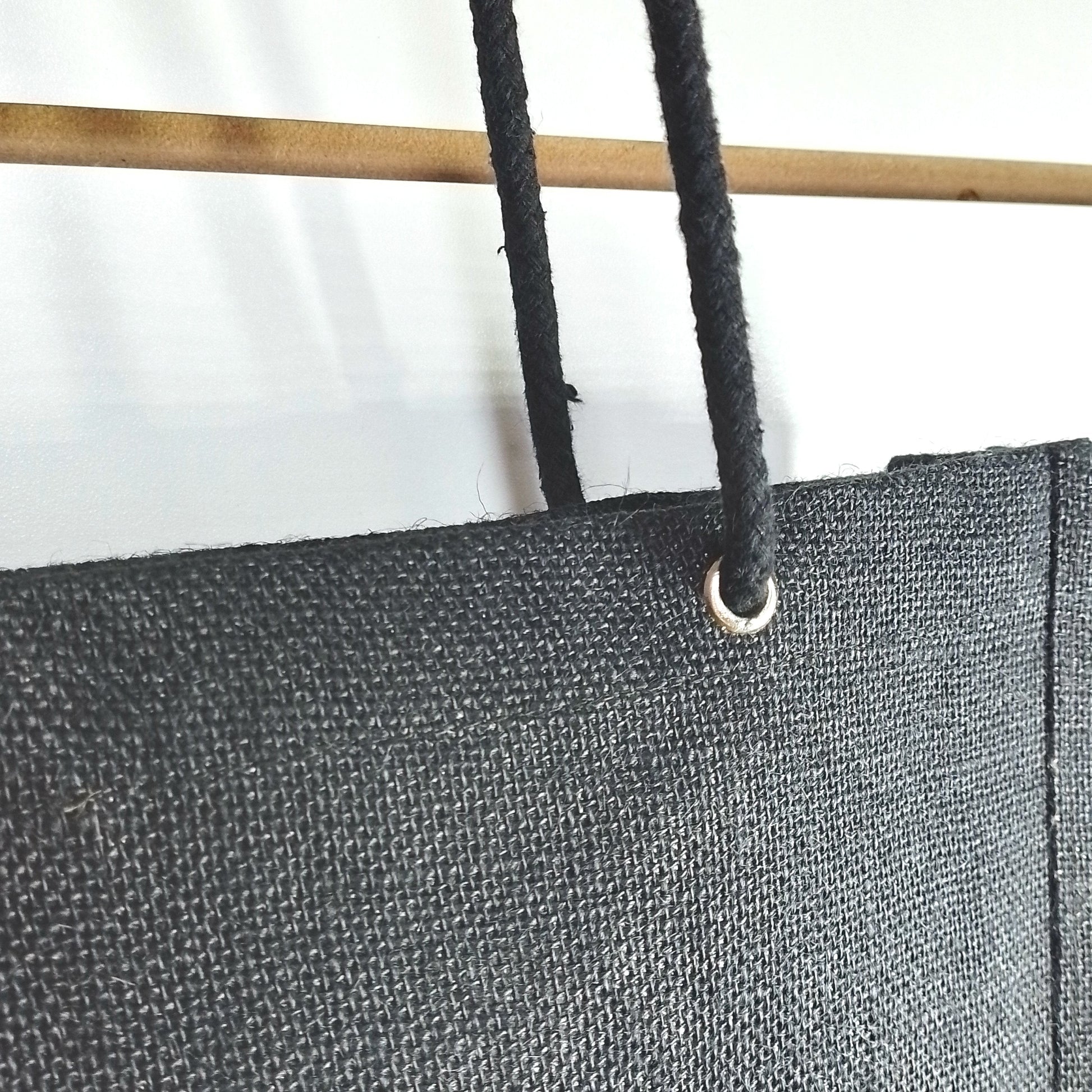 Close-up of a black canvas or heavy fabric panel with a metal grommet and black cord, shown against a white background with part of a wooden pole visible.