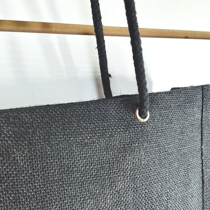 Close-up of a black canvas or heavy fabric panel with a metal grommet and black cord, shown against a white background with part of a wooden pole visible.