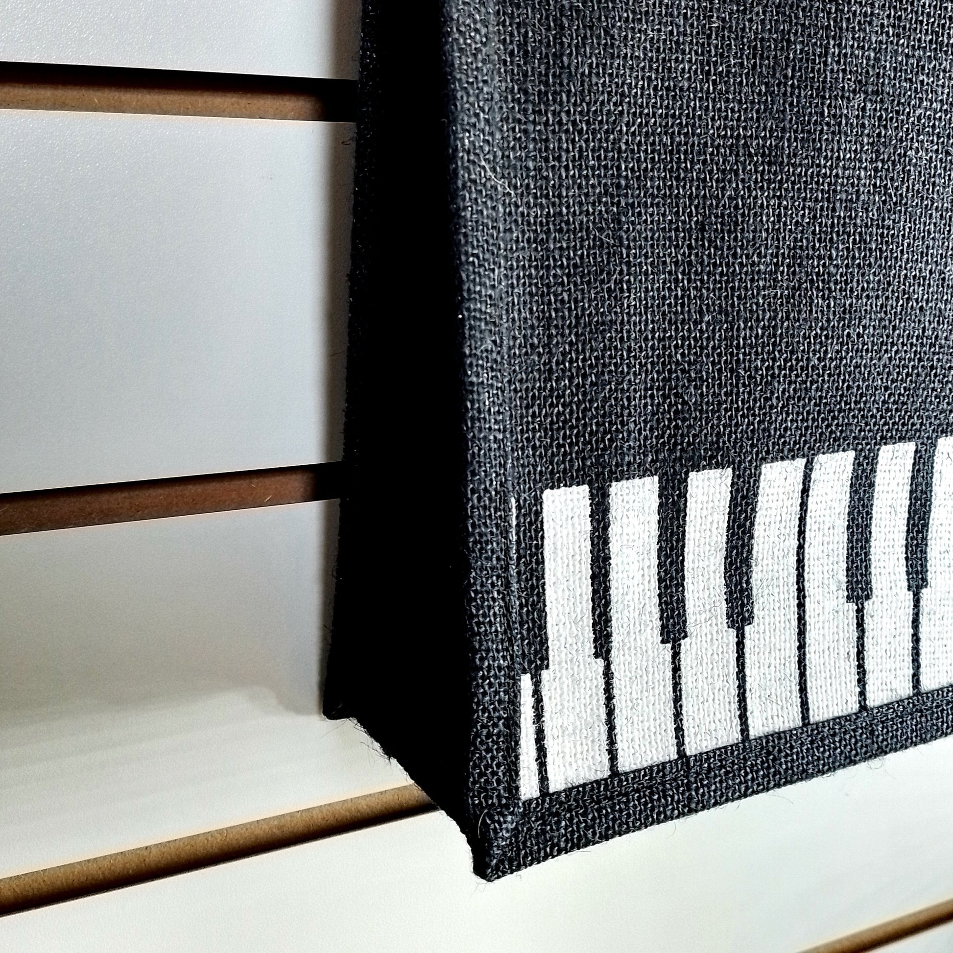 A close-up of a black woven fabric item, likely a basket or bag, featuring a white piano-key pattern on one side, set against a white slatted wall background.