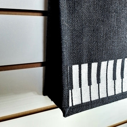 A close-up of a black woven fabric item, likely a basket or bag, featuring a white piano-key pattern on one side, set against a white slatted wall background.