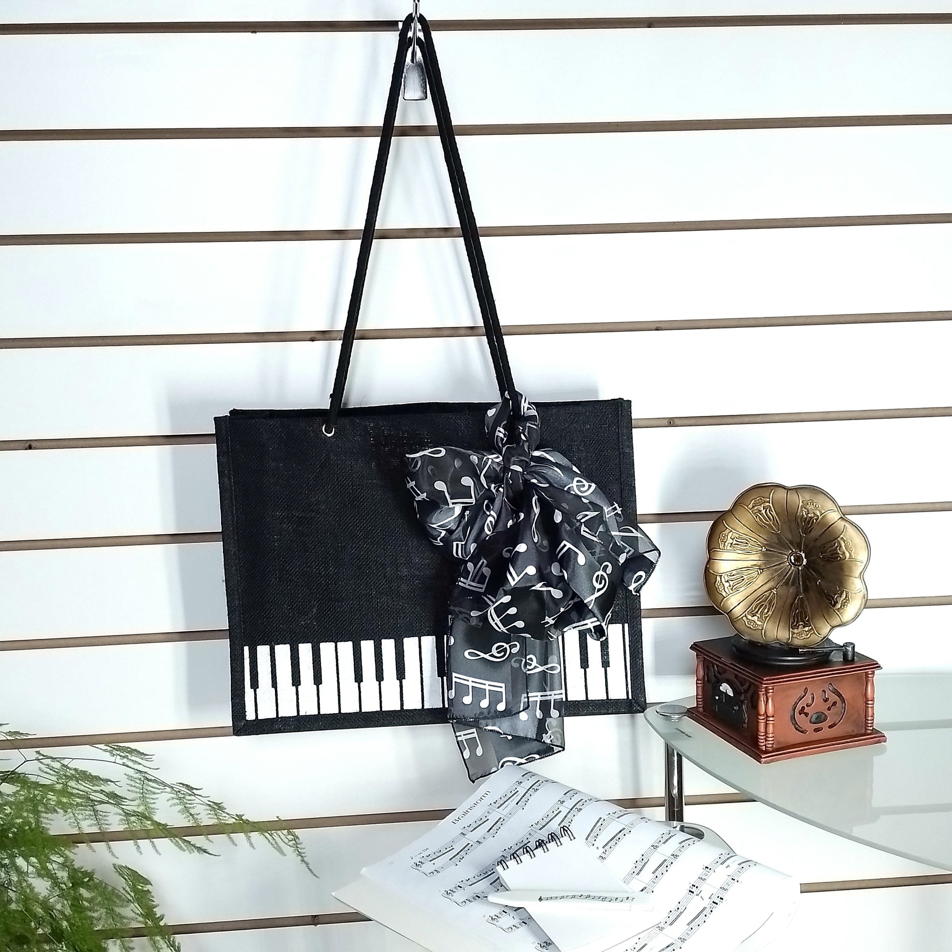 A black Music Themed Reusable Shopping Bag, styled with a black and white music note print scarf, displayed on a white slatted wall alongside sheet music, a small wooden box, and a gold gramophone ornament.
