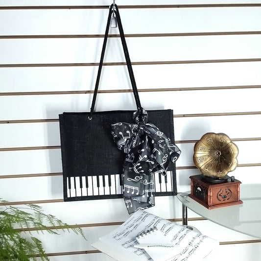 A black Music Themed Reusable Shopping Bag, styled with a black and white music note print scarf, displayed on a white slatted wall alongside sheet music, a small wooden box, and a gold gramophone ornament.