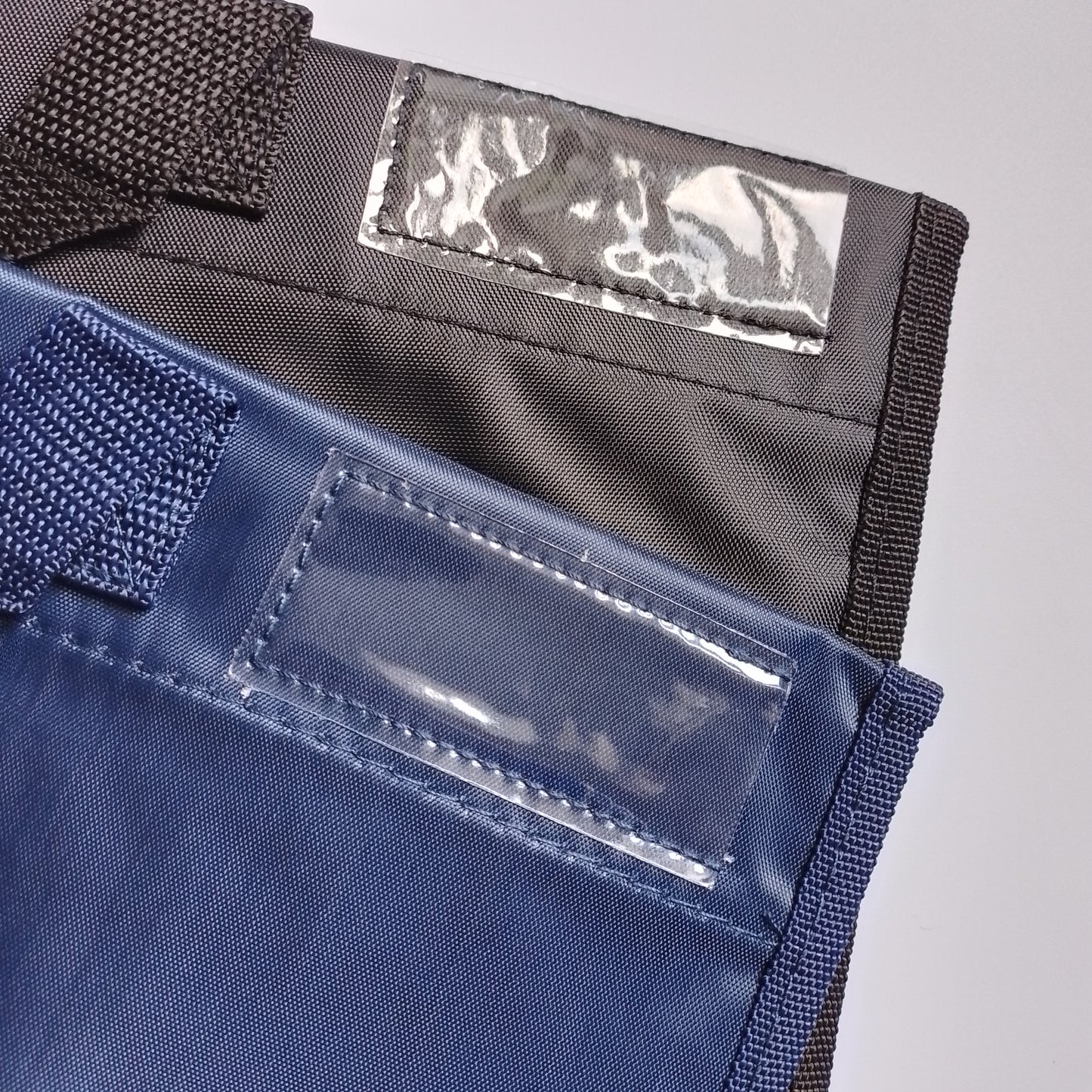 Close-up of two fabric pouches or sleeves with clear plastic ID windows and dark woven straps, shown on a light gray background.