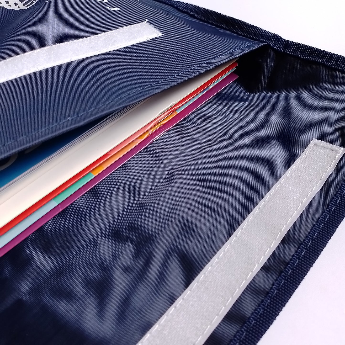 Close-up of a navy blue fabric music bag with a flap, showing a stack of colourful paper or card inserts and a reflective light grey strip on the inside.