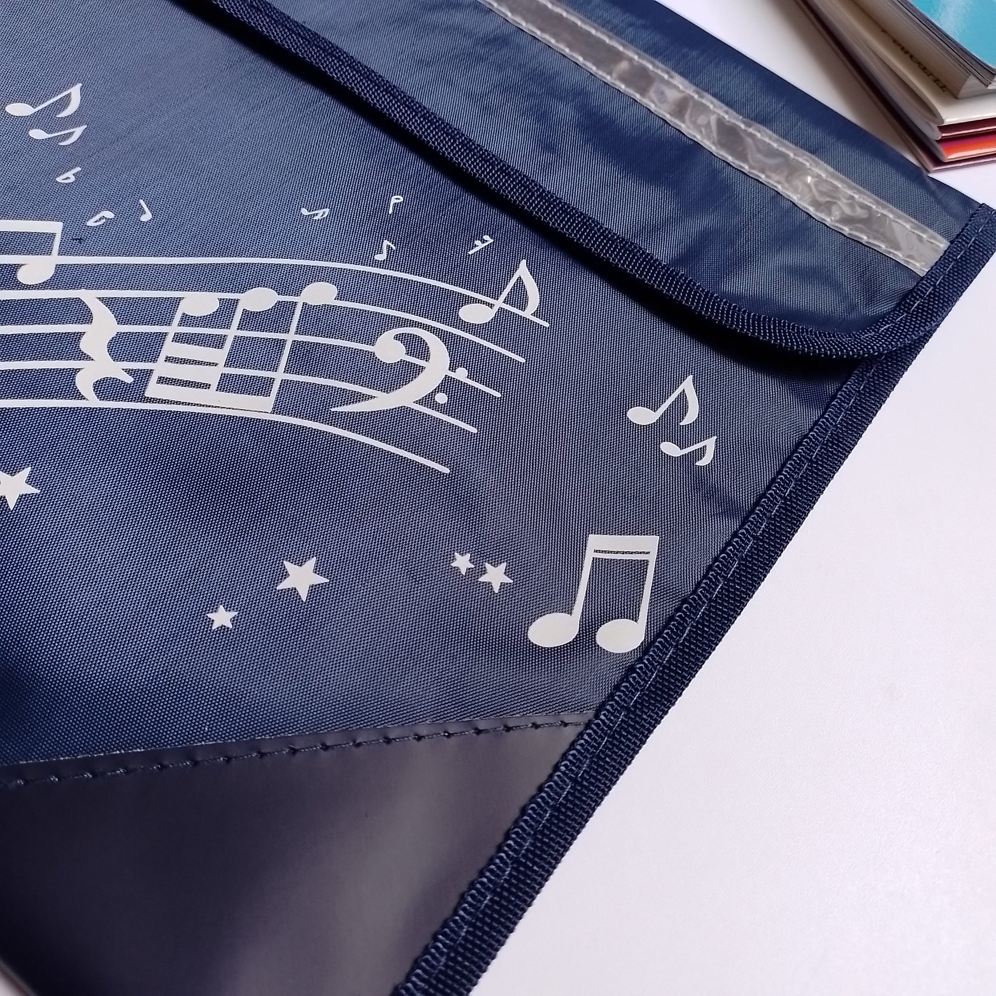 A navy blue fabric music bag with white musical notes, staff lines, and stars printed on it, shown on a white surface with part of a book visible in the top right corner.