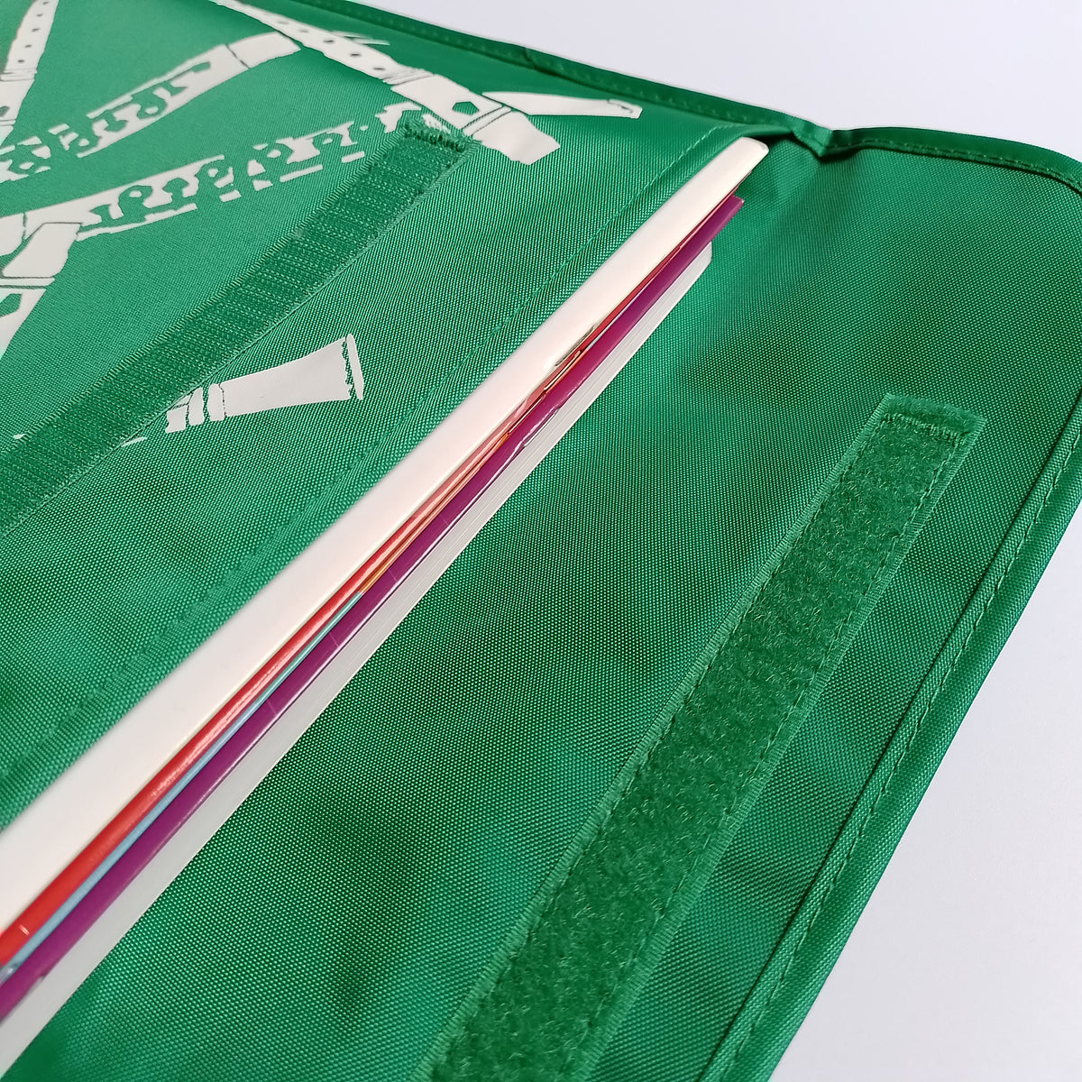 Close-up of a green nylon bag with a long strip of Velcro closure. The school bag contains a stack of colourful books.