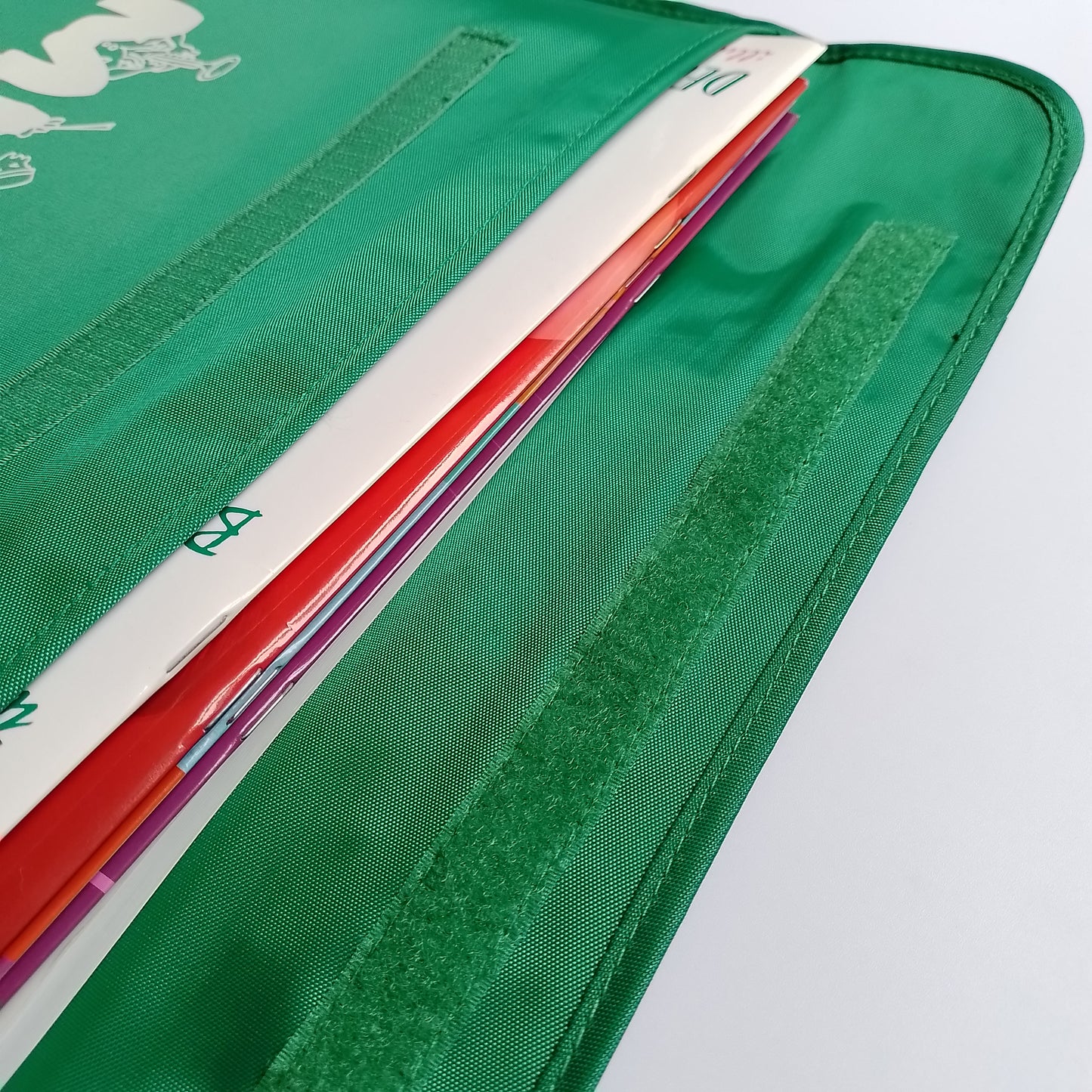 Close-up of a green nylon bag with a long strip of Velcro closure. The school bag contains a stack of colourful books.