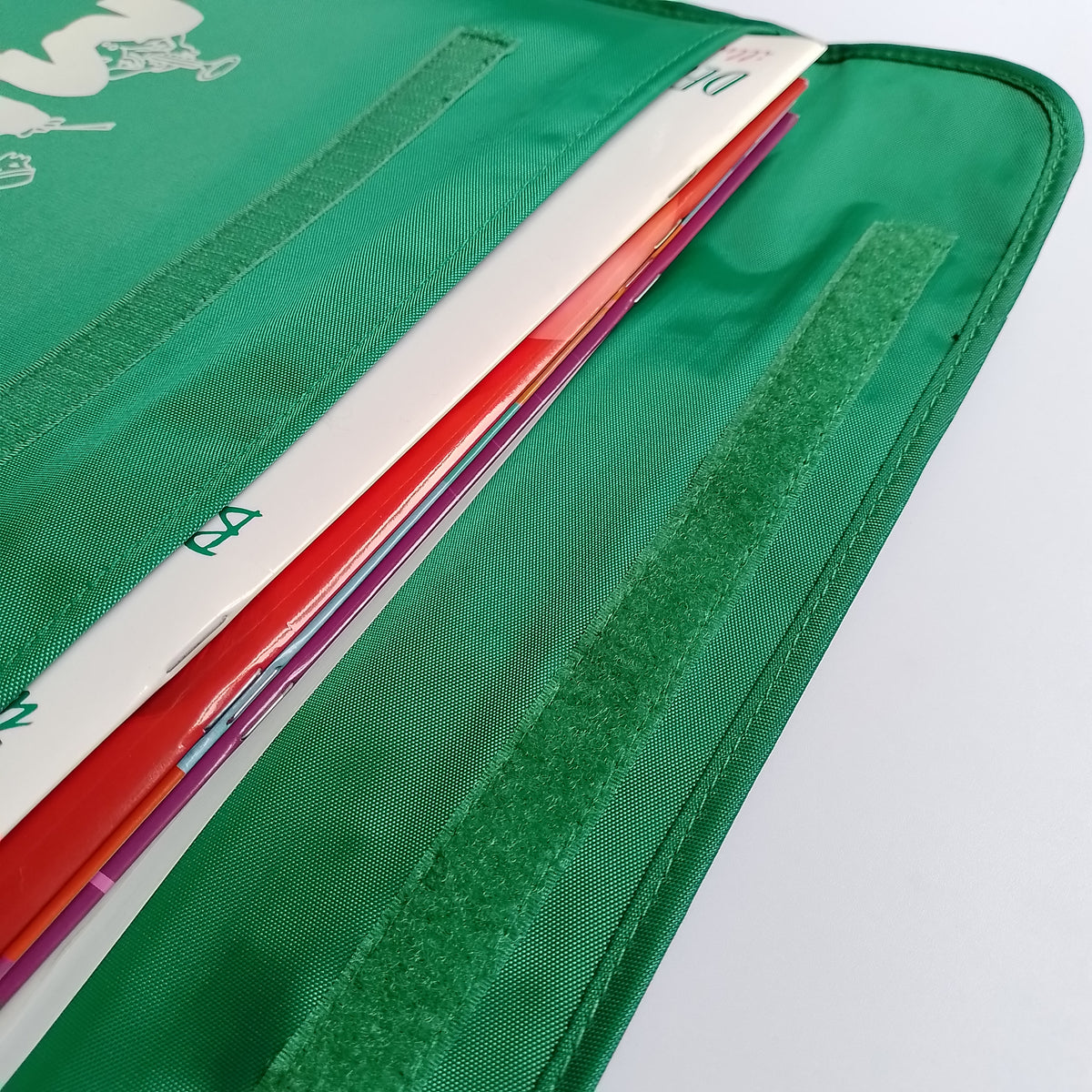 Close-up of a green nylon bag with a long strip of Velcro closure. The school bag contains a stack of colourful books.