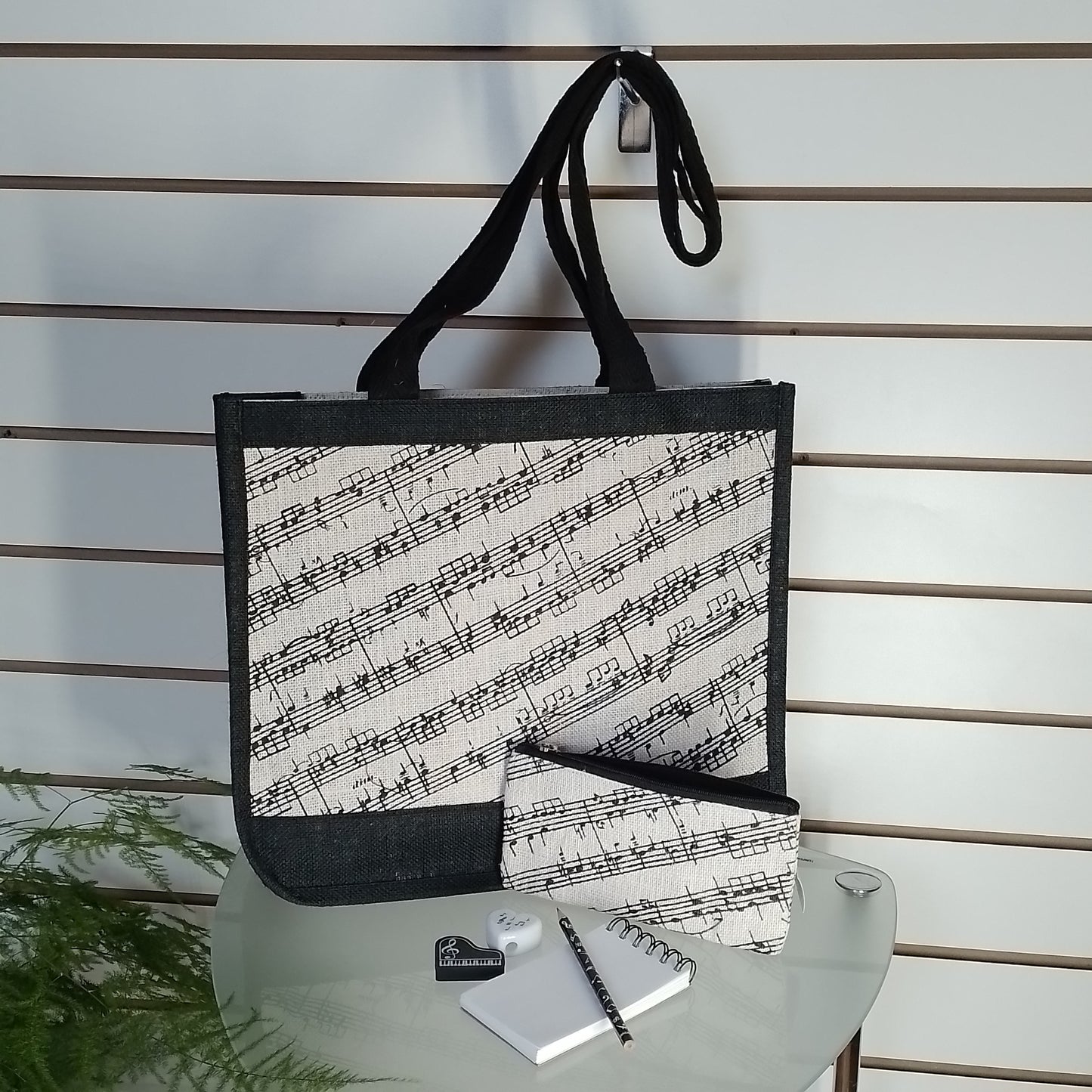 A tote bag with a black and white musical staff pattern, displayed on a glass table with a matching pouch, notebook, and pen against a slatwall background.