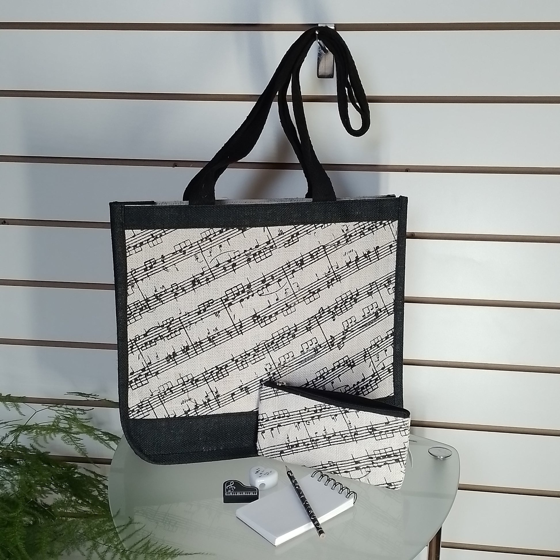 A tote bag with a black and white musical staff pattern, displayed on a glass table with a matching pouch, notebook, and pen against a slatwall background.