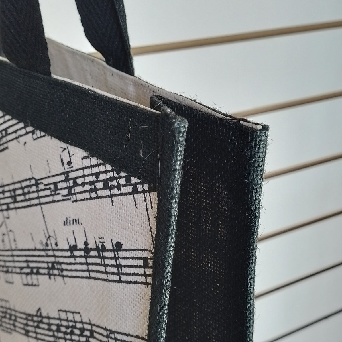 Close-up of a tote bag corner showing a black-and-white musical sheet music print on a canvas-like fabric, with black woven trim and handles against a white slatted background.