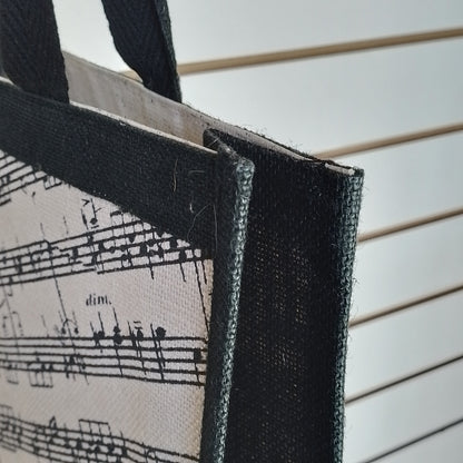 Close-up of a tote bag corner showing a black-and-white musical sheet music print on a canvas-like fabric, with black woven trim and handles against a white slatted background.