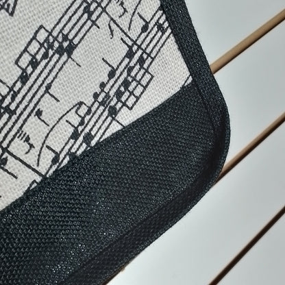 Close-up of a square textile product corner with a black mesh border and a white fabric section printed with black musical notes and staff lines, resting on a light surface with thin wooden slats visible.