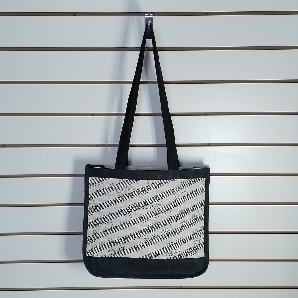 A canvas tote bag with black handles and a black-and-white musical sheet music print, hanging from a metal hook on a white slatwall.