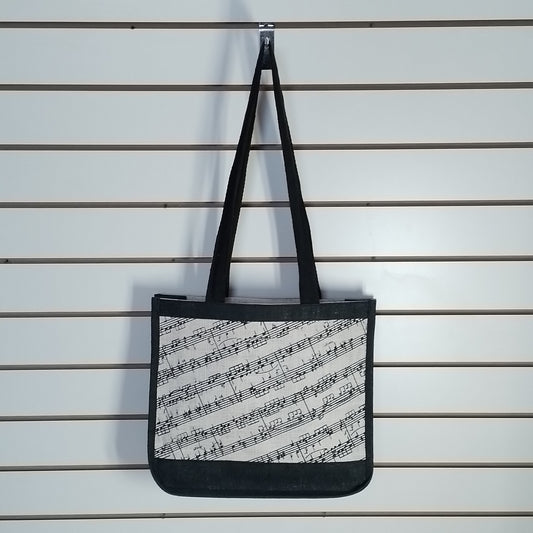 A canvas tote bag with black handles and a black-and-white musical sheet music print, hanging from a metal hook on a white slatwall.