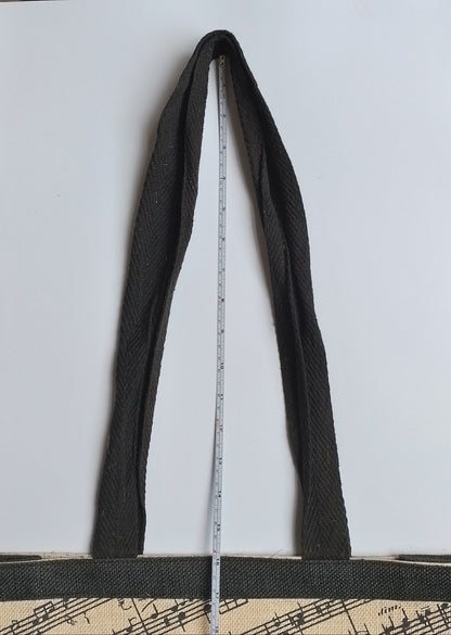 A black woven tote bag with a beige printed pattern at the bottom, shown with a metal tape measure hanging from its strap to indicate size, set against a plain white background.