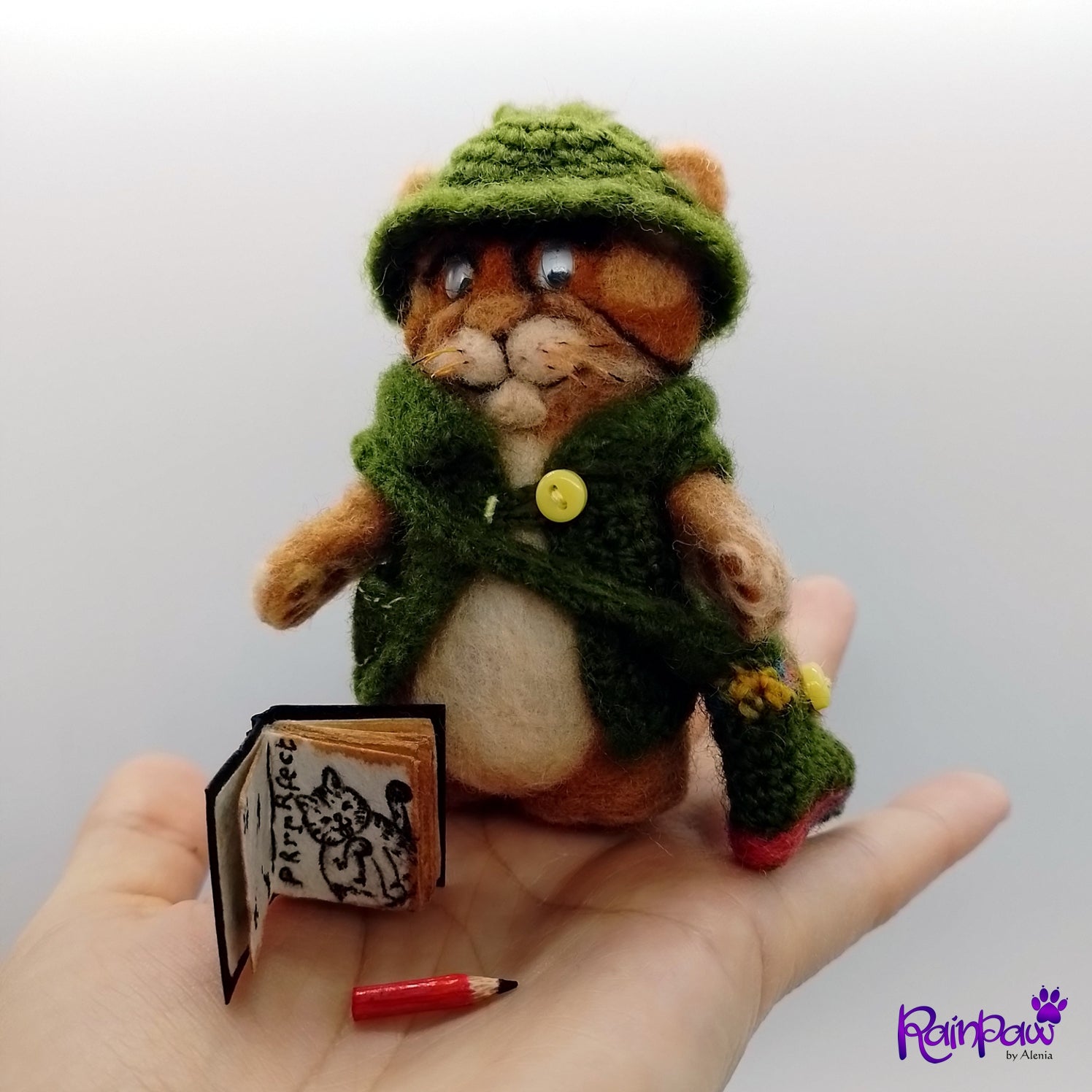 Small stuffed ginger cat/tiger with a green outfit held in a hand, with a miniature book and pencil on a white background.